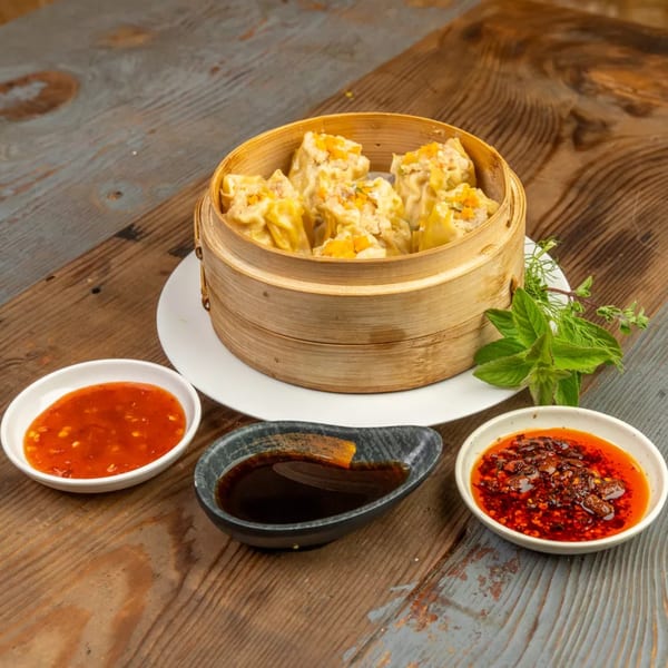 Steamed dumplings in a bamboo steamer, with three dipping sauces and fresh herbs on a wooden table.