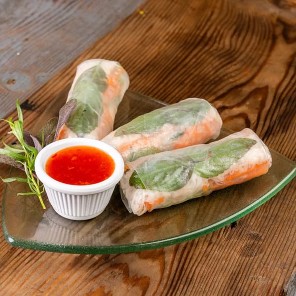 Three fresh spring rolls with sweet chili dipping sauce and herbs on a glass plate.