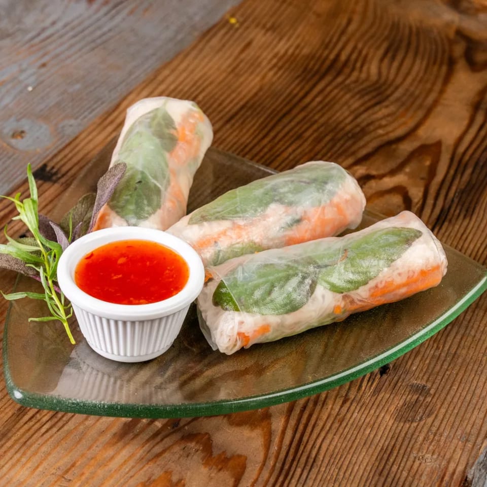 Three fresh spring rolls with sweet chili dipping sauce and herbs on a glass plate.