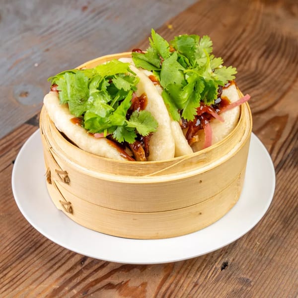Steamed bao buns with cilantro, sauce, and pickled onions in a bamboo steamer on a white plate.