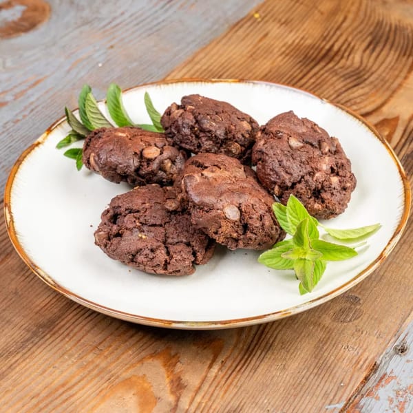 Five dark chocolate cookies with nuts and mint leaves on a white plate.