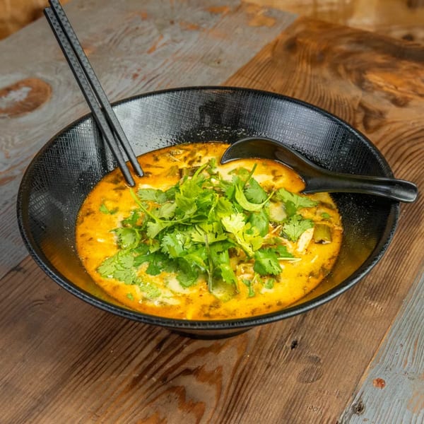 A black bowl of vibrant yellow curry soup with green cilantro, chopsticks, and a spoon on a wooden table.